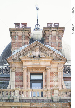 An ornate brick and stone facade showcases elegant architecture centered around a doorway framed by classic fluted columns. Above the entryway, a triangular pediment encloses a detailed stone bust An ornate brick and stone facade showcases elegant architecture centered around a doorway framed by classic fluted columns. Above the entryway, a triangular pediment encloses a detailed stone bust 139902690