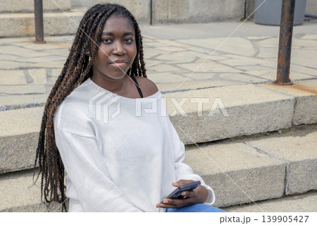 Young African student sitting on steps with smartphone, urban campus 139905427