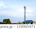 Telecommunication tower standing tall in a residential neighborhood under a bright blue sky. High steel lattice mast with cellular antennas near house roofs and green trees. 5G network concept. 139905971