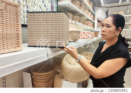 Asian middle-aged woman shopping for woven storage baskets in a modern home goods store. Female customer choosing eco-friendly rattan boxes on shelves. Retail shopping and home decoration. Asian middle-aged woman shopping for woven storage baskets in a modern home goods store. Female customer choosing eco-friendly rattan boxes on shelves. Retail shopping and home decoration. 139905991