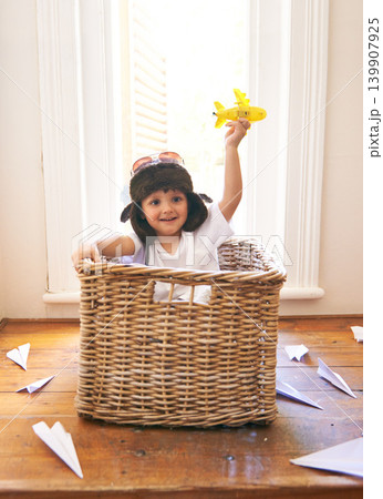 Child, smile and airplane toy in home, playful and freedom of youth with paper for fun and games. Little kid, happiness and hand in air with body in basket with hat on head as helmet with imagination 139907925