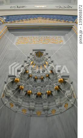 A close up of a marble mihrab shows detailed muqarnas designs and gold leaf accents. Soft natural light highlights the features in a mosque setting. A close up of a marble mihrab shows detailed muqarnas designs and gold leaf accents. Soft natural light highlights the features in a mosque setting. 139908872