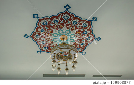 A detailed view shows a ceiling in a mosque, featuring a geometric star pattern in red and blue. A brass chandelier hangs below the ornate design. A detailed view shows a ceiling in a mosque, featuring a geometric star pattern in red and blue. A brass chandelier hangs below the ornate design. 139908877
