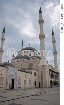 The scene shows a large mosque complex with tall minarets and a wide courtyard. People walk around the area while clouds cover the sky. The architecture reflects historical design. The scene shows a large mosque complex with tall minarets and a wide courtyard. People walk around the area while clouds cover the sky. The architecture reflects historical design. 139908881
