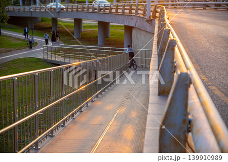 Cyclist riding down a ramp under a modern bridge. Man on bicycle descending metal path in city urban park. People walking on trail during sunset. Infrastructure and commute concept. Cyclist riding down a ramp under a modern bridge. Man on bicycle descending metal path in city urban park. People walking on trail during sunset. Infrastructure and commute concept. 139910989