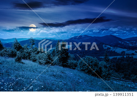 mysterious field on hillside in mountains near village at night. full moon over carpathian mountain landscape in summer. countryside view of alpine fields and rolling hills. cloudy sky 139911351