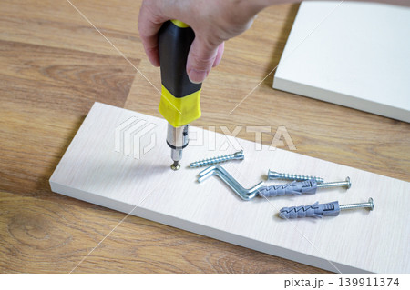 Hand screws a self-tapping screw into a furniture panel on a laminate floor. Fasteners, dowels and hex keys are ready for DIY home repairs work. 139911374