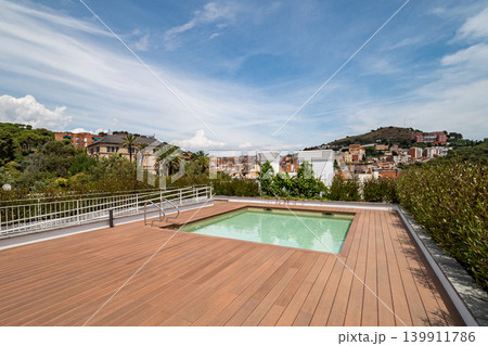 A modern rooftop terrace with a swimming pool. The area features wooden decking and overlooks a scenic view of buildings and hills under a clear blue sky. A modern rooftop terrace with a swimming pool. The area features wooden decking and overlooks a scenic view of buildings and hills under a clear blue sky. 139911786