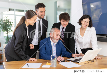 Friendly group of businesspeople working together, everyone looking at vacation photos on laptop by mature man and laughing in corporate office 139914379