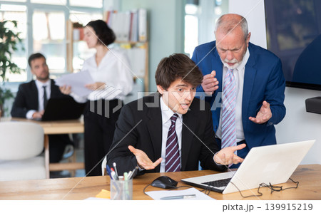 Elderly man scolding young male colleague working on laptop Elderly man scolding young male colleague working on laptop 139915219