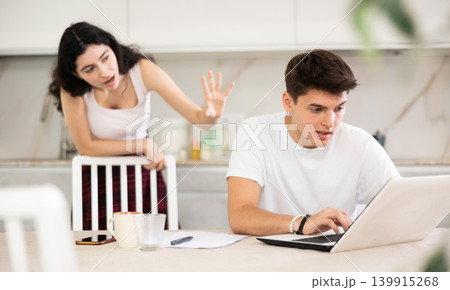 Sad freelancer husband works at home on laptop and listens to her wife reproaches in frustration. Sad freelancer husband works at home on laptop and listens to her wife reproaches in frustration. 139915268