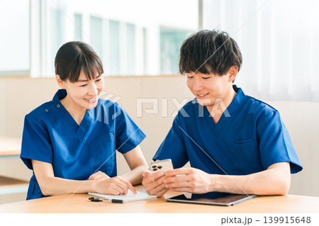 スマホを見ながら会話する先輩看護師と若い男性看護師 スマホを見ながら会話する先輩看護師と若い男性看護師 139915648