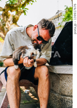 A caucasian 30 years old young man sitting on a stone bench and hugging a cute fluffy cat outdoors A caucasian 30 years old young man sitting on a stone bench and hugging a cute fluffy cat outdoors 139917676