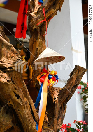 Traditional vietnamese conical hat non la ornament hanging with colorful ribbons on an ancient decorative tree trunk at a temple courtyard Traditional vietnamese conical hat non la ornament hanging with colorful ribbons on an ancient decorative tree trunk at a temple courtyard 139917702