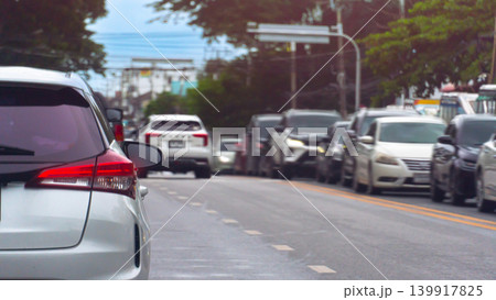 Vehicles are lined up in a heavy daytime traffic jam on a wide city road. A silver car is prominent in the foreground lane, with lush green trees lining the roadside in the background. 139917825