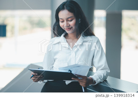 Professional woman reviewing business documents on a clipboard while leaning in a modern office, expressing focused analysis, report checking, organized workflow, and everyday corporate productivity. 139918314