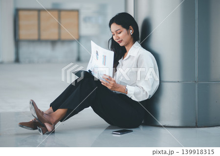 Professional woman sitting on the office floor reading a business report, showing focused analysis, independent work, document review, quiet concentration, and workplace productivity. Professional woman sitting on the office floor reading a business report, showing focused analysis, independent work, document review, quiet concentration, and workplace productivity. 139918315