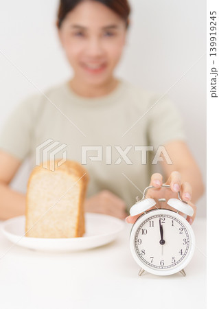 Happy smiling woman with whole wheat bread and alarm clock. concept of intermittent fasting, IF, meal timing, and choosing healthy carbohydrates for a balanced diet and weight loss management Happy smiling woman with whole wheat bread and alarm clock. concept of intermittent fasting, IF, meal timing, and choosing healthy carbohydrates for a balanced diet and weight loss management 139919245