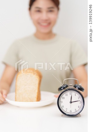 Happy smiling woman with whole wheat bread and alarm clock. concept of intermittent fasting, IF, meal timing, and choosing healthy carbohydrates for a balanced diet and weight loss management Happy smiling woman with whole wheat bread and alarm clock. concept of intermittent fasting, IF, meal timing, and choosing healthy carbohydrates for a balanced diet and weight loss management 139919246