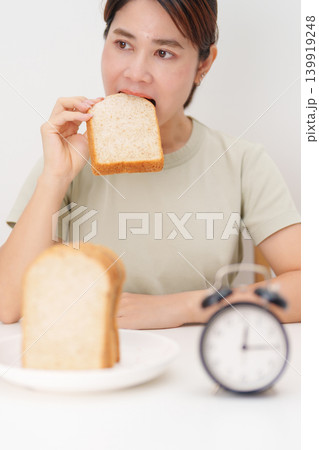 Happy smiling woman eating whole wheat bread with alarm clock. concept of intermittent fasting, IF, meal timing, and choosing healthy carbohydrates for a balanced diet and weight loss management Happy smiling woman eating whole wheat bread with alarm clock. concept of intermittent fasting, IF, meal timing, and choosing healthy carbohydrates for a balanced diet and weight loss management 139919248