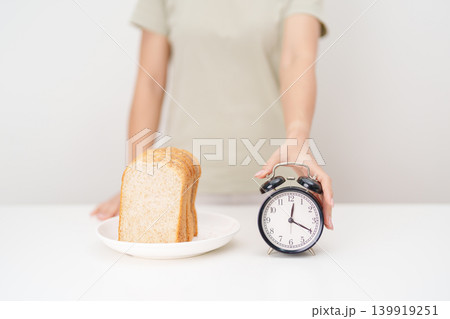 Happy smiling woman with whole wheat bread and alarm clock. concept of intermittent fasting, IF, meal timing, and choosing healthy carbohydrates for a balanced diet and weight loss management Happy smiling woman with whole wheat bread and alarm clock. concept of intermittent fasting, IF, meal timing, and choosing healthy carbohydrates for a balanced diet and weight loss management 139919251