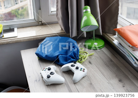 Two gaming controllers sit on a wooden desk next to a green lamp and stacks of books. Two gaming controllers sit on a wooden desk next to a green lamp and stacks of books. 139923756