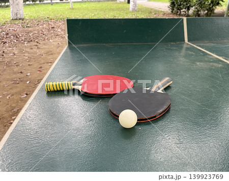 Two table tennis paddles and a ball are placed on a green table in a park during the day Two table tennis paddles and a ball are placed on a green table in a park during the day 139923769