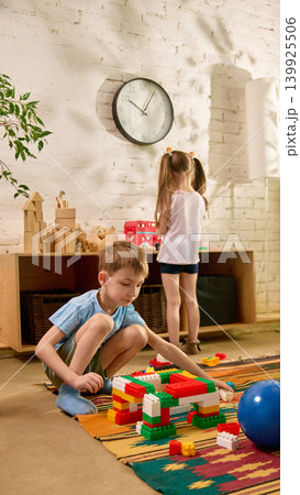 Boy constructing colorful block bridge while girl plays near shelf in bright room. Boy constructing colorful block bridge while girl plays near shelf in bright room. 139925506