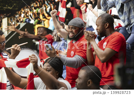 Happy soccer fan checking phone after last minute winning bet during intense football match filled with cheering supporters and crowd excitement. Celebrating a successful cash out and gambling win. 139928723