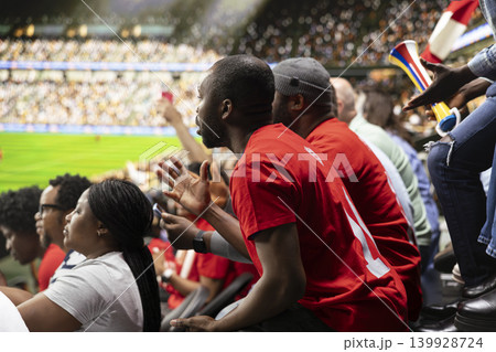 Focused football supporters watching dramatic final minutes of live soccer match from crowded stadium stands with red scarves. Sports fans asking for the final whistle in the stadium. 139928724