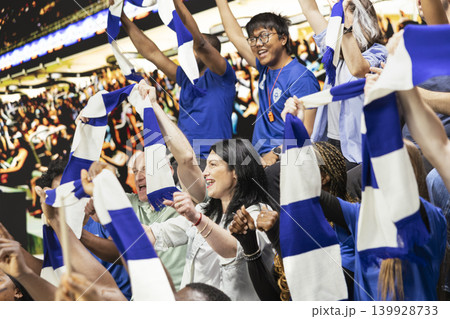 Singing blue team football fans waving scarves and cheering together in packed stadium stands during energetic live soccer match. Team spirit and celebration inside the packed stadium. 139928733