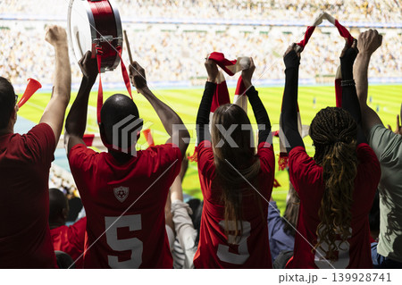 Back view of passionate supporters cheering in the stadium stands during football match filled with crowd noise and excitement. Diverse soccer ultras wave raised scarves, play fan drums. 139928741