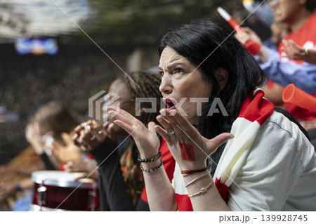 Emotional mature female football fan reacting with open hands in disbelief during crucial soccer match moment from stadium stands. Dejected supporters of the losing team remain stunned. Emotional mature female football fan reacting with open hands in disbelief during crucial soccer match moment from stadium stands. Dejected supporters of the losing team remain stunned. 139928745