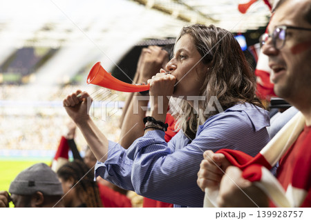 Passionate female football fan blowing a red vuvuzela while cheering in crowded stadium stands during an intense live soccer match. Diverse multigenerational supporters in the fan section. 139928757