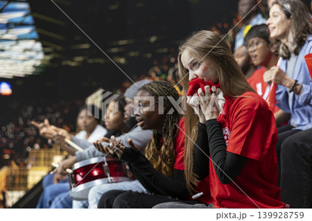 Anxious young football supporter biting red scarf in crowded stadium stands during tense live game, modern football with women empowerment and inclusion. Suspense and anticipation. 139928759