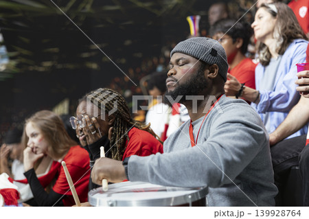 Black football supporter with drum closing eyes in stadium stands during tense soccer match, surrounded by anxious red fans. Emotional silence during crucial moment in the last minute. Black football supporter with drum closing eyes in stadium stands during tense soccer match, surrounded by anxious red fans. Emotional silence during crucial moment in the last minute. 139928764