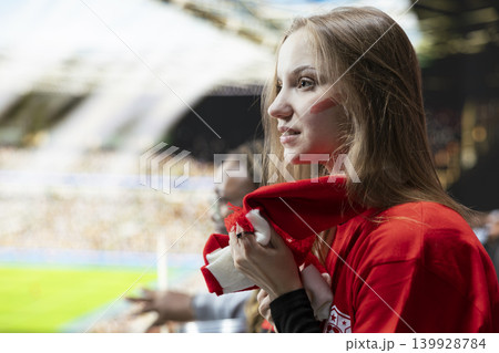 Side profile of young woman football supporter holding red scarf in tension during soccer game from stadium stands. Tense crowd showing anxiety and suspense, inclusive football community. Side profile of young woman football supporter holding red scarf in tension during soccer game from stadium stands. Tense crowd showing anxiety and suspense, inclusive football community. 139928784