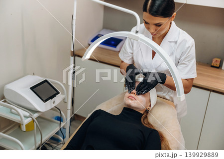 White cream, quality product. Female cosmetologist performing a facial skincare procedure using a cosmetic device in a beauty clinic White cream, quality product. Female cosmetologist performing a facial skincare procedure using a cosmetic device in a beauty clinic 139929889