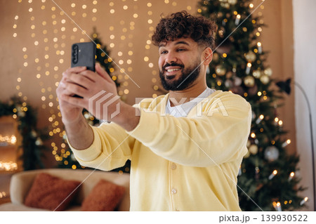Making a selfie by using smartphone. Man posing in front of a Christmas tree in a festive home setting Making a selfie by using smartphone. Man posing in front of a Christmas tree in a festive home setting 139930522