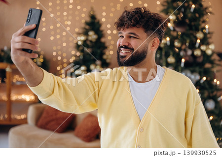 Making a selfie by using smartphone. Man posing in front of a Christmas tree in a festive home setting 139930525