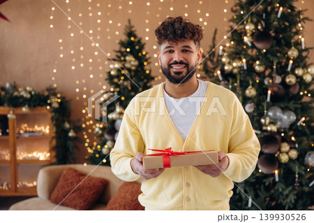 Gift box is in hands. Man posing in front of a Christmas tree in a festive home setting Gift box is in hands. Man posing in front of a Christmas tree in a festive home setting 139930526