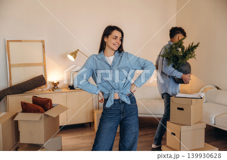 Hands are on the waist, woman is standing. Positive couple placing a potted plant in their new apartment during the moving process Hands are on the waist, woman is standing. Positive couple placing a potted plant in their new apartment during the moving process 139930628