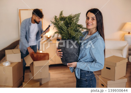 Looking at the camera and smiling. Positive couple placing a potted plant in their new apartment during the moving process Looking at the camera and smiling. Positive couple placing a potted plant in their new apartment during the moving process 139930644