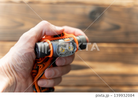 Hand holding an orange and gray handheld flashlight with dual LED lights against a wooden background, showcasing its design and functionality for outdoor use 139932084