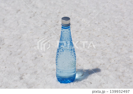 Blue glass water bottle standing on a textured white salt crust. Concept of absolute purity, refreshing hydration and natural mineral source. Blue glass water bottle standing on a textured white salt crust. Concept of absolute purity, refreshing hydration and natural mineral source. 139932497