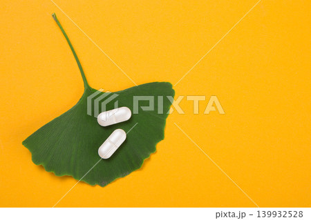 Ginkgo biloba green leaf and two white medicine capsules on bright orange background. Herbal supplement for brain health and memory concept. Ginkgo biloba green leaf and two white medicine capsules on bright orange background. Herbal supplement for brain health and memory concept. 139932528