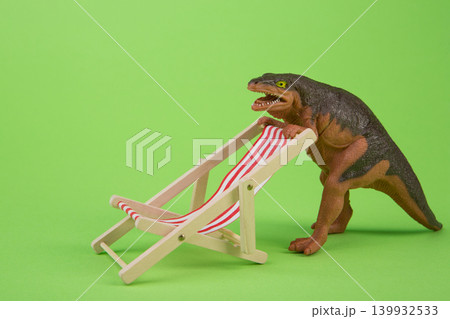Funny toy dinosaur adjusting a miniature beach deck chair on a bright green background. Funny toy dinosaur adjusting a miniature beach deck chair on a bright green background. 139932533