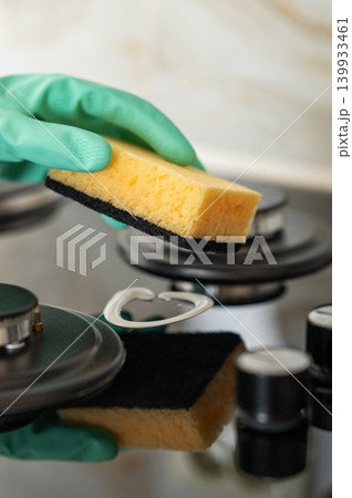 A close-up of a person's hand in a rubber glove cleaning a gas cooktop with a sponge. A close-up of a person's hand in a rubber glove cleaning a gas cooktop with a sponge. 139933461