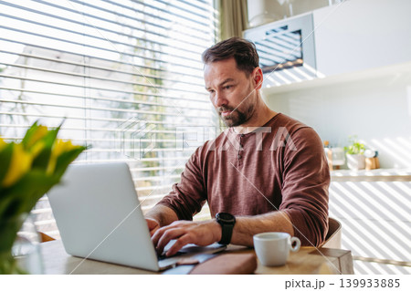 Man sitting in kitchen working remotely on laptop. 139933885