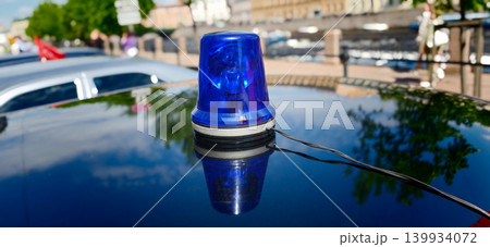Blue emergency light on top of a black vehicle reflecting in sunlight, with trees and buildings visible in the background along a city street Blue emergency light on top of a black vehicle reflecting in sunlight, with trees and buildings visible in the background along a city street 139934072